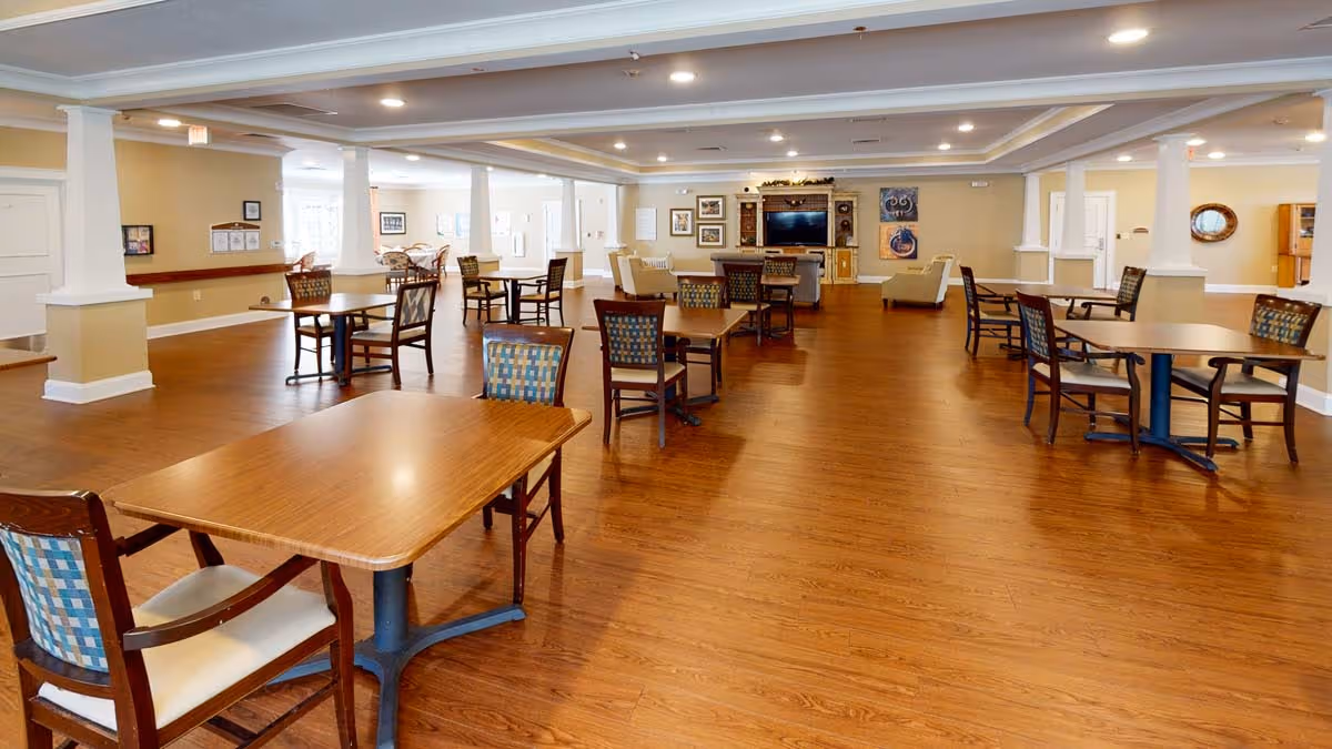 Spacious dining/activity room with multiple wooden tables and chairs and a TV seating area at the far end.