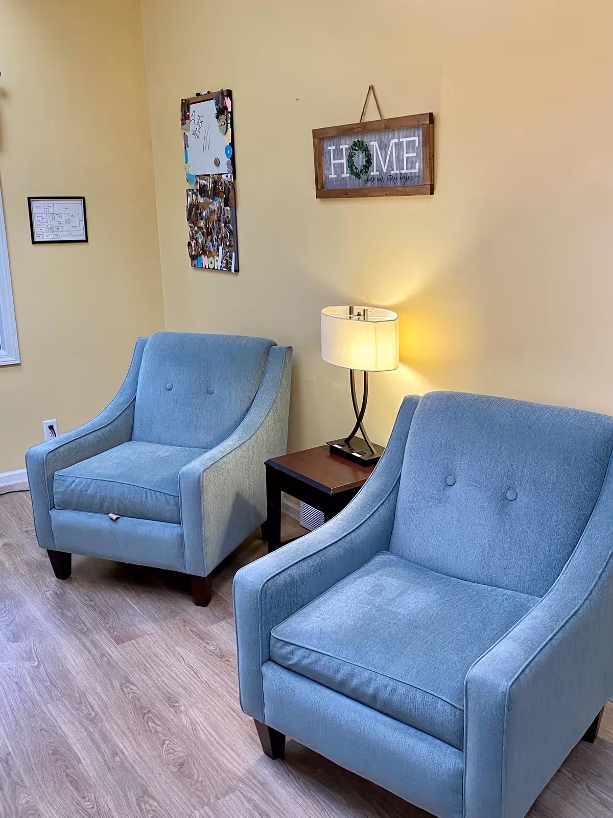 Two light blue upholstered armchairs with button tufting are placed on a light wood floor against a pale yellow wall. Between the chairs is a small dark wood side table with a modern lamp that has a white shade. On the wall above the table is a wooden framed sign that reads 'HOME' with a wreath replacing the letter O. To the left on the wall is a bulletin board with photos and notes, and a framed floor plan is partially visible.
