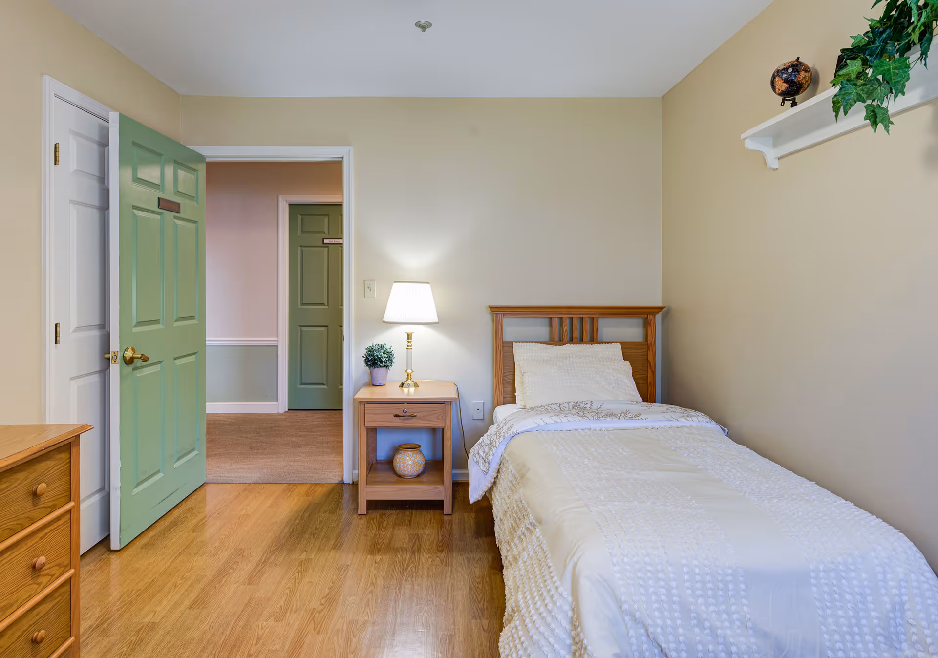 A simple bedroom with a single wooden bed covered with a white textured blanket and pillow. Next to the bed is a wooden nightstand with a small lamp and a potted plant. The room has light beige walls and a wooden floor. There is a green door open to a hallway with another green door visible. A white shelf with a small globe and a green plant is mounted on the wall above the bed.