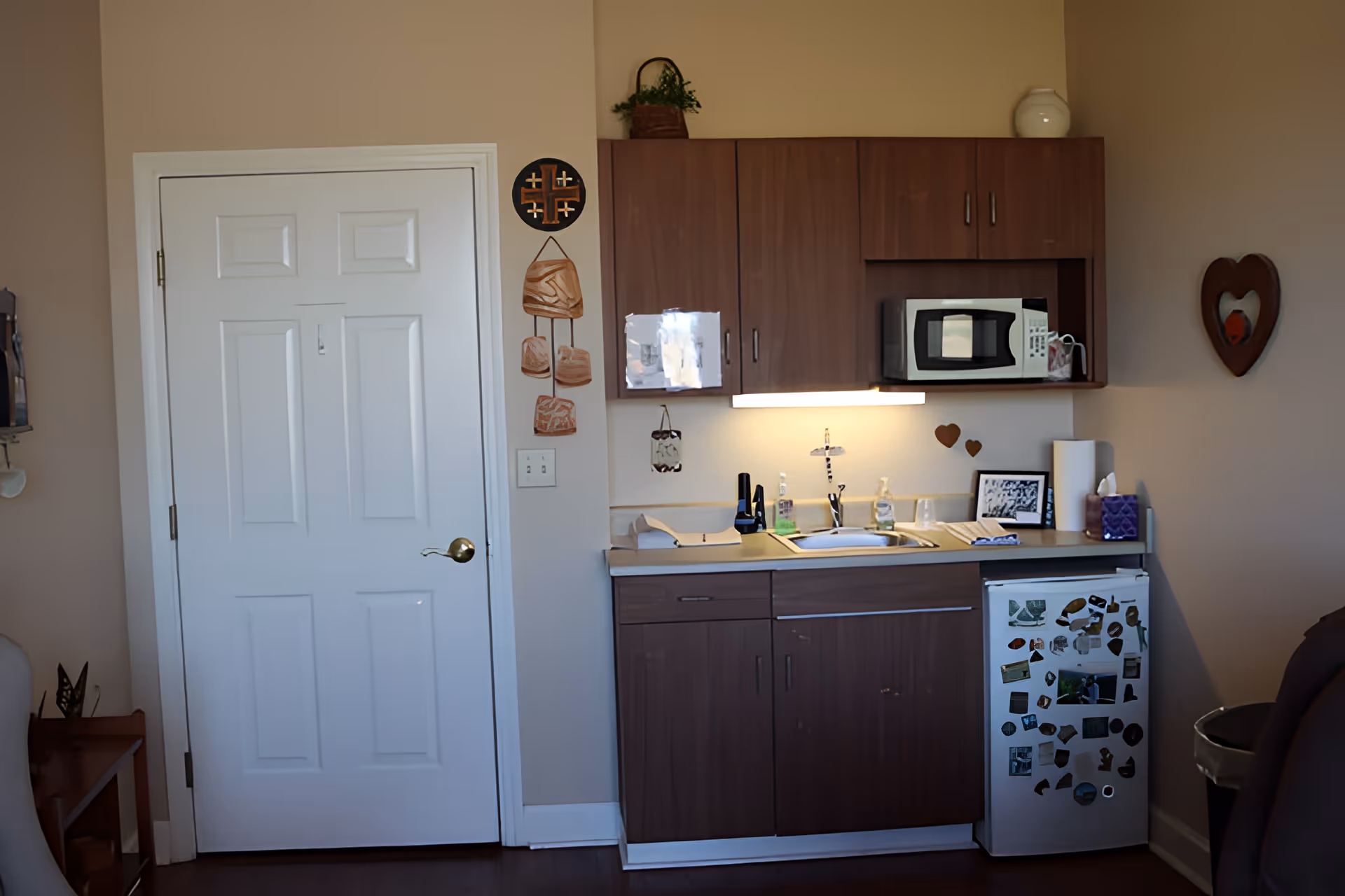 Small kitchenette area with wooden cabinets, a white mini refrigerator covered with magnets, a microwave on the countertop, a sink with soap dispensers, and various decorative items on the walls and countertop. A closed white door is to the left of the kitchenette.