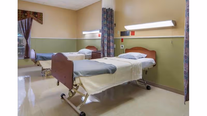 A room with two hospital-style beds separated by a privacy curtain. Each bed has a pillow and blanket, with a window covered by curtains on the left side. The walls are painted beige and green, and there are medical equipment panels above each bed.
