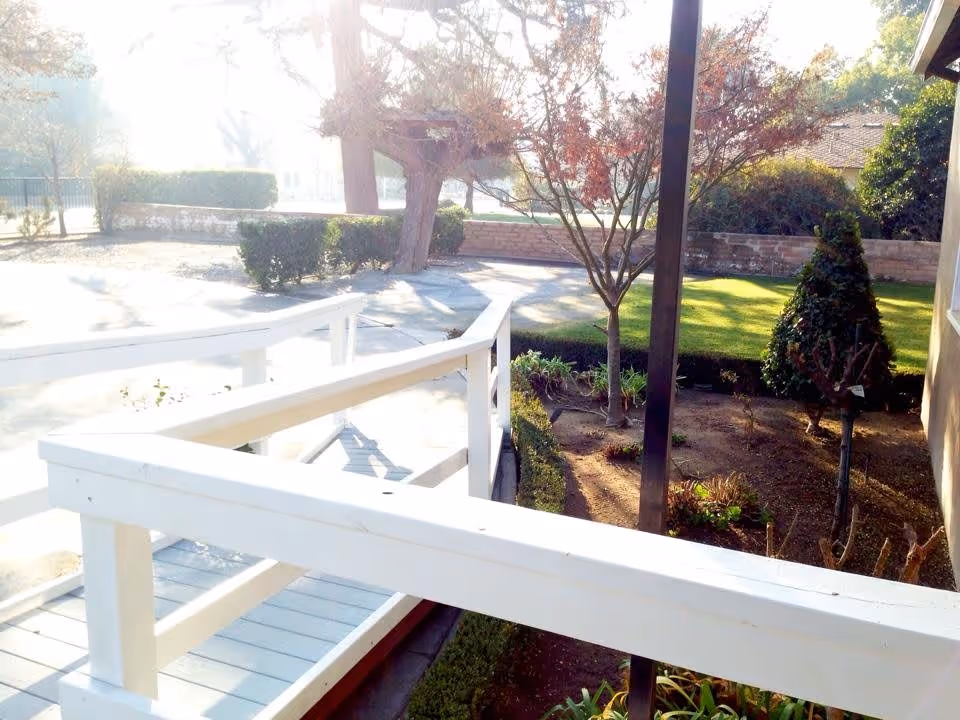 Outdoor view of a garden area with trimmed bushes, trees, and a white wooden ramp railing in the foreground. Sunlight brightens the scene, casting shadows on the ground and highlighting the greenery.