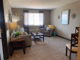 Cozy living room with a sofa, armchair, coffee table, side table with lamp, and a window with curtains.