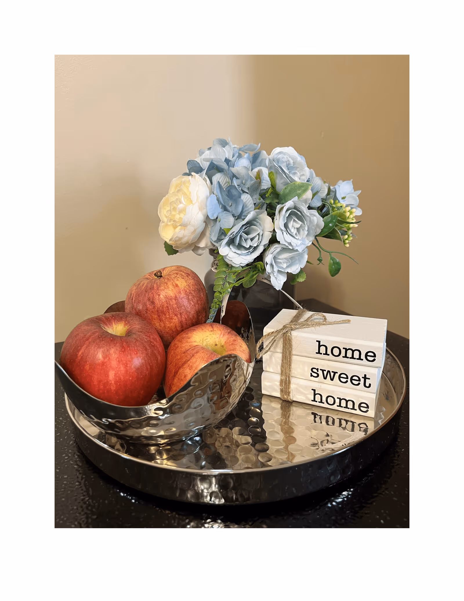 A decorative arrangement on a hammered metal tray featuring a hammered metal bowl with three red apples, a small bouquet of light blue and white artificial flowers, and a stack of three white wooden blocks tied with twine that read 'home sweet home'.