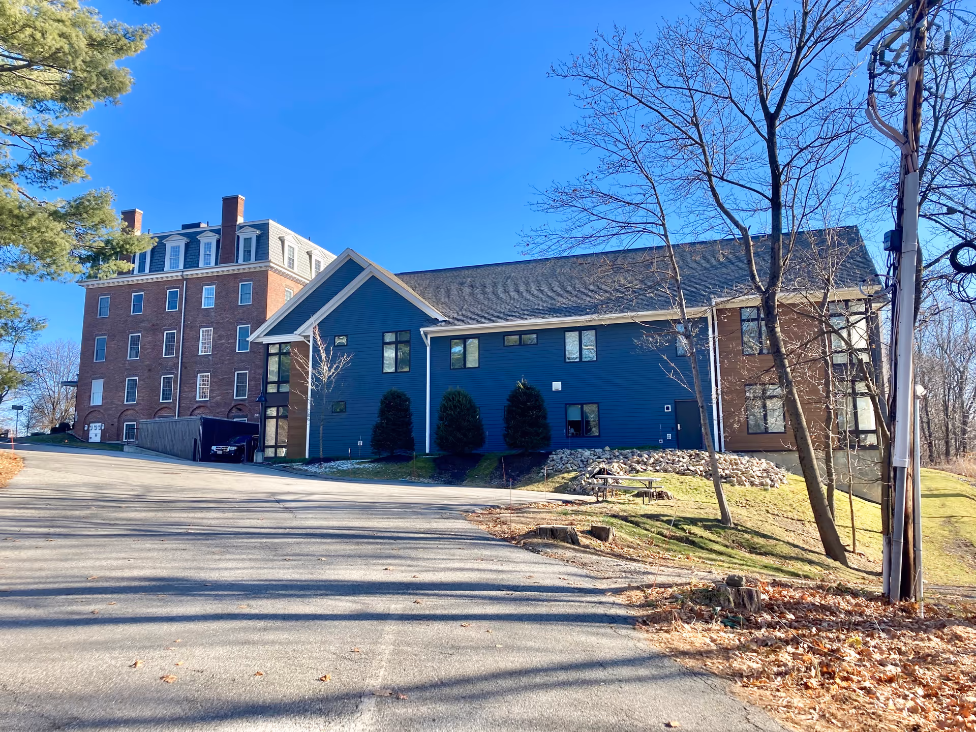 Exterior view of a senior living facility with two connected buildings, one brick and one blue with large windows, surrounded by trees and a clear blue sky.