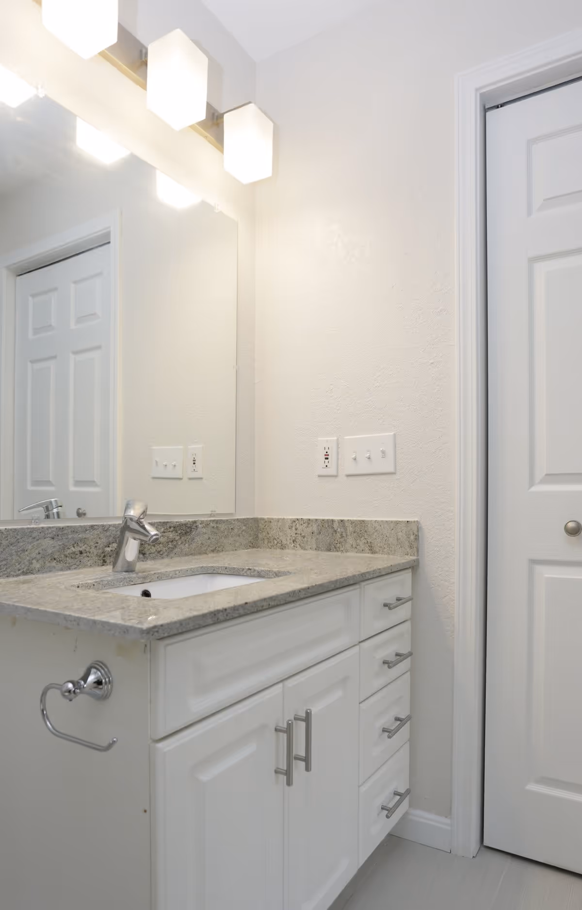A clean bathroom vanity with a granite countertop, an under-mount sink, a modern chrome faucet, white cabinetry with silver handles, a large mirror above the sink, three square light fixtures above the mirror, and a closed white door to the right.