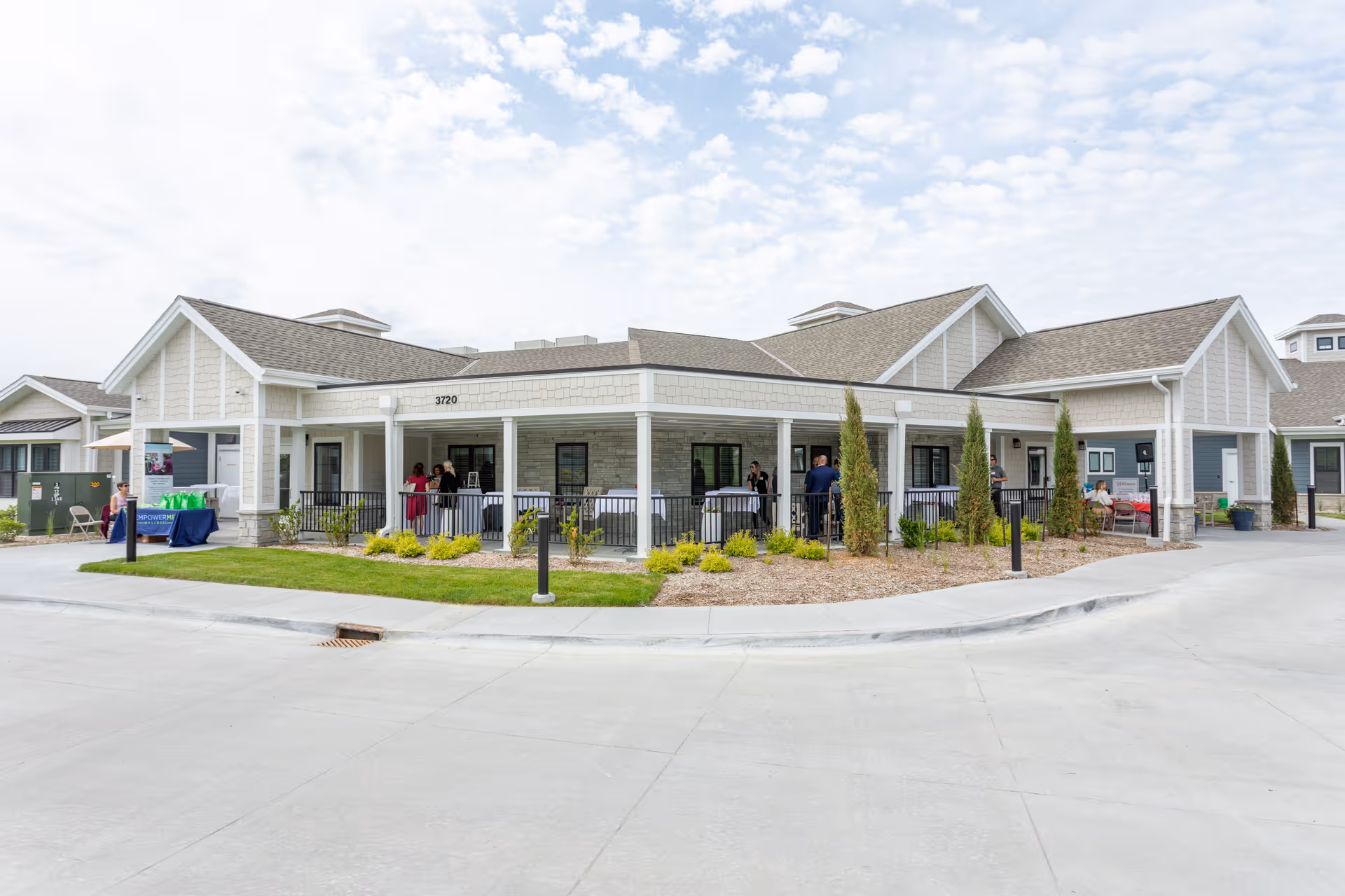 Front exterior of a single-story senior living building with a covered porch, people on the patio, and landscaped grounds.