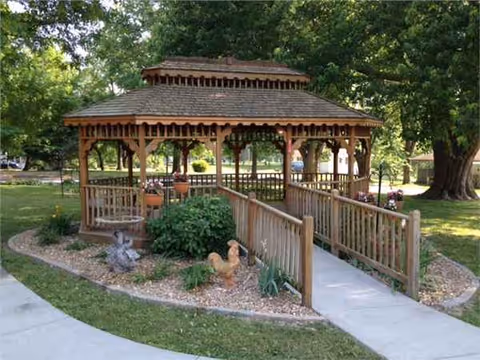 A wooden gazebo with benches sits in a landscaped park area near trees.