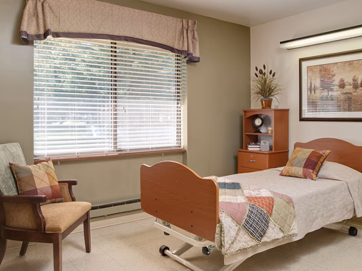 A single bed with a wooden headboard and footboard, covered with a quilt and a decorative pillow, is positioned next to a wooden nightstand with shelves and a clock. There is a cushioned armchair with a patterned pillow near a large window with blinds and a valance. A framed landscape painting hangs on the wall above the bed.