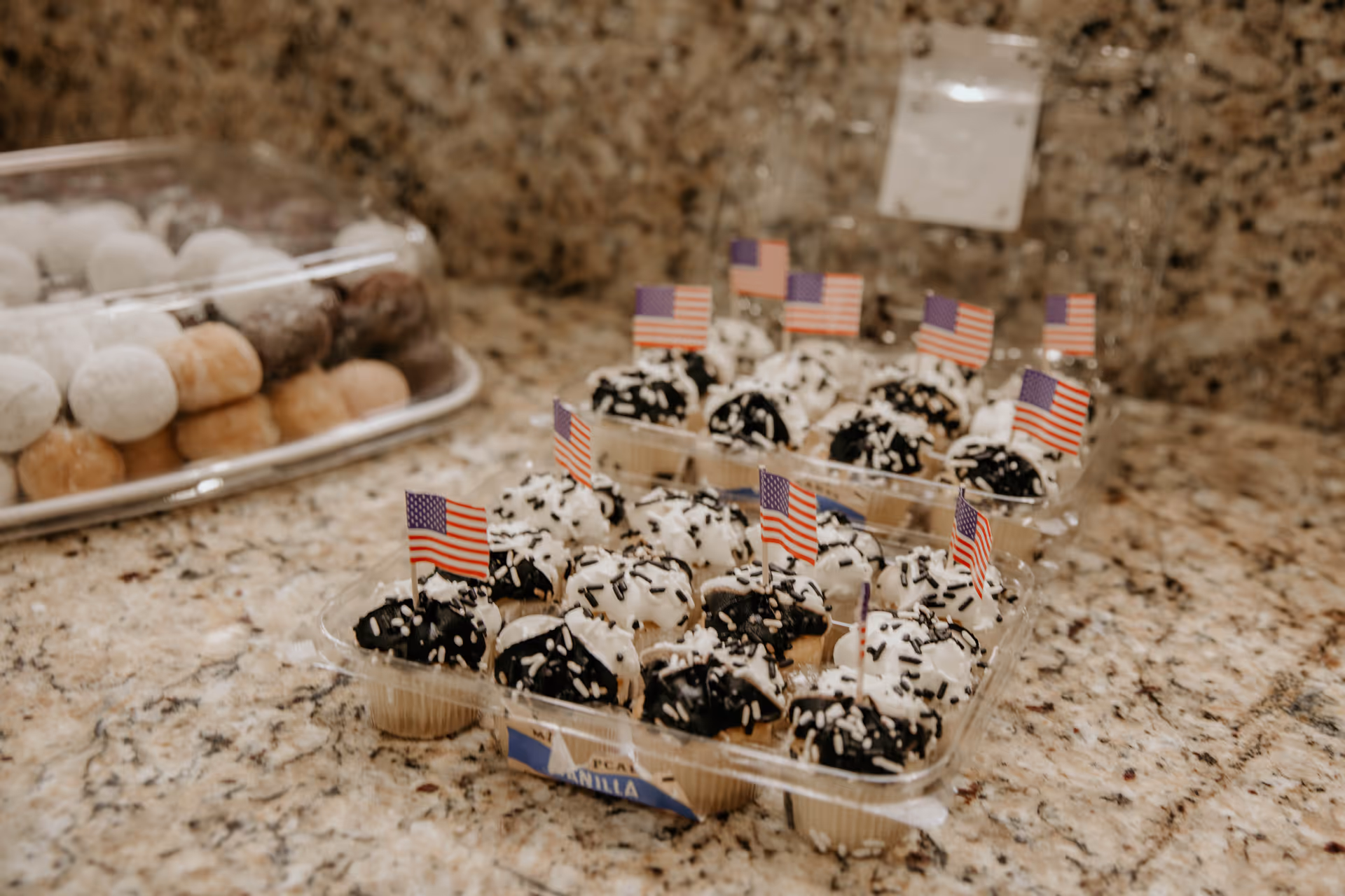 Close-up view of mini cupcakes with white and chocolate frosting topped with small American flag toothpicks, placed on a granite countertop. In the background, there is a clear plastic container with powdered sugar-coated donut holes.