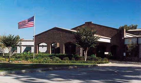 Exterior of The Oaks Nursing and Rehabilitation, a brick senior care facility with flag and trees.