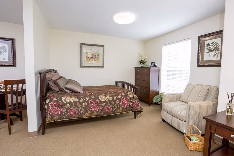 A cozy bedroom in a senior living facility featuring a bed with a floral bedspread, several pillows, a wooden dresser, a striped armchair, and framed artwork on the walls. A window with blinds allows natural light into the room. Part of a dining area with a wooden chair and table is visible to the left.