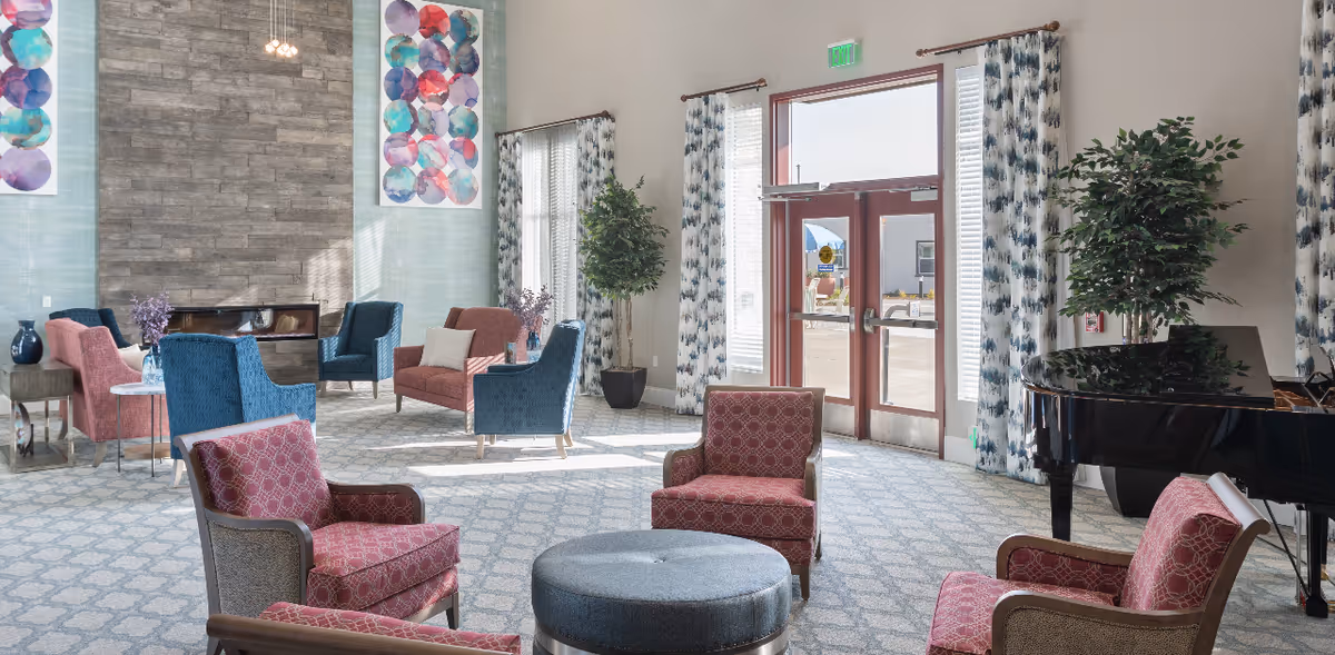 A spacious and well-lit living room area with multiple upholstered chairs and sofas arranged around a central round ottoman. The room features a large stone fireplace, colorful abstract wall art, tall windows with patterned curtains, potted plants, and a grand piano near the entrance door.