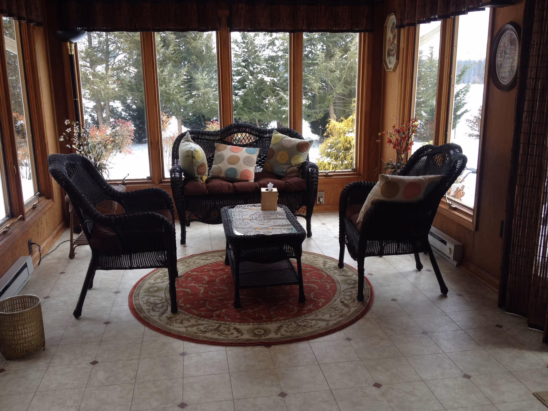 A cozy sunroom with large windows showing a snowy outdoor scene. The room contains black wicker furniture including a loveseat and two chairs with cushions and colorful polka dot pillows. A small wicker coffee table with a lace doily and tissue box sits on a round red and beige patterned rug. There are decorative plants near the windows and wood-paneled walls.