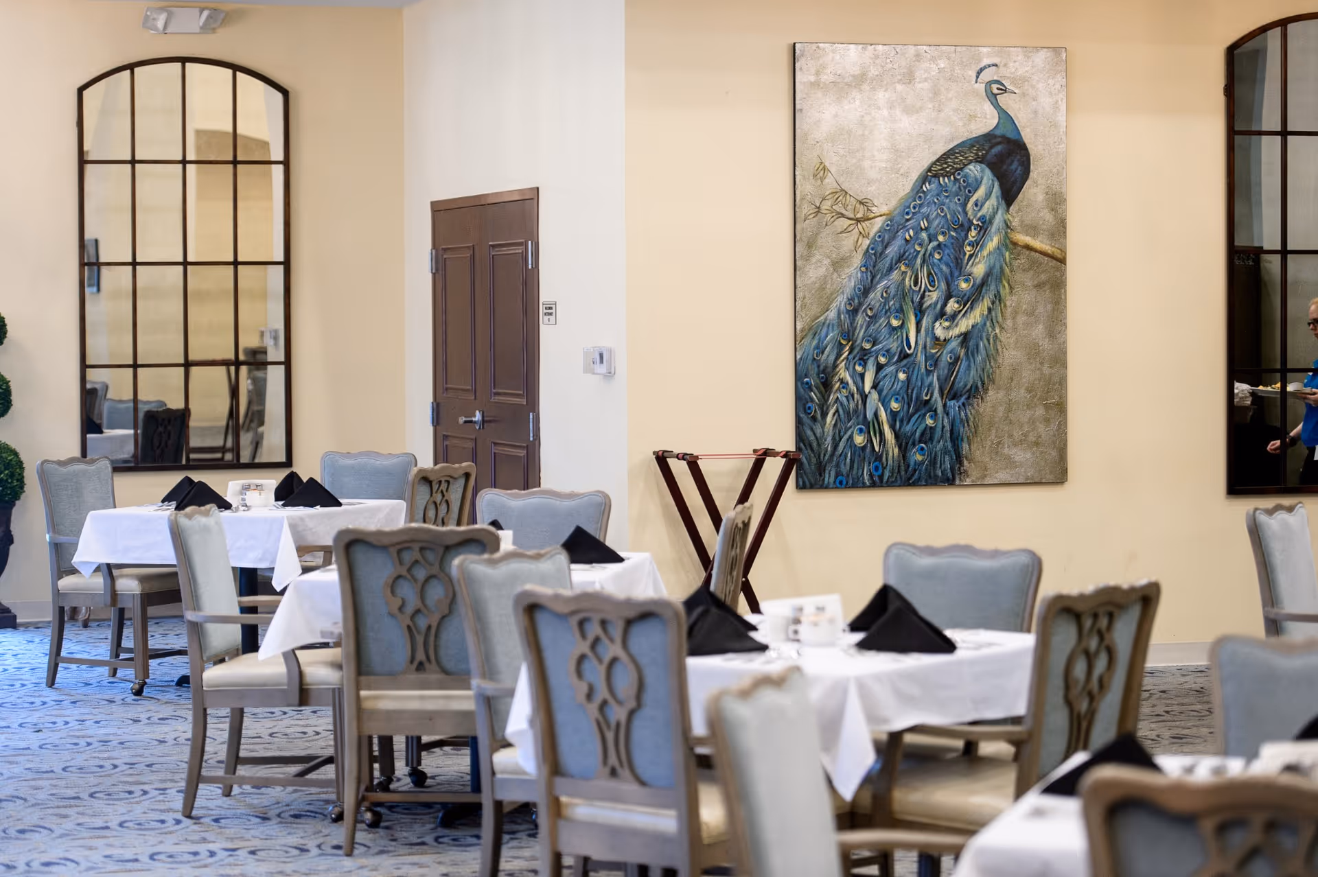 Dining room with tables covered in white tablecloths and black napkins, surrounded by upholstered chairs. A large painting of a peacock hangs on the wall, and there are two large arched mirrors reflecting parts of the room. A person is partially visible on the right side of the image.