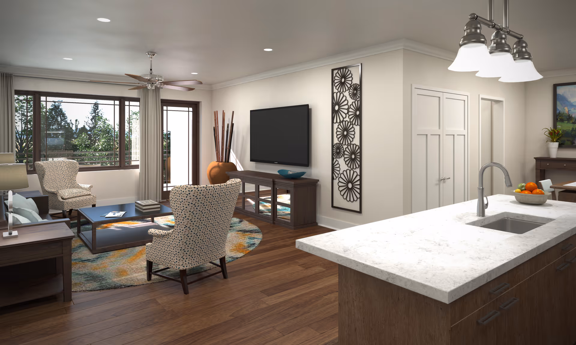 A bright and modern living room area with wooden flooring, two patterned armchairs, a coffee table on a colorful round rug, a flat-screen TV mounted on the wall above a wooden console, and large windows letting in natural light. To the right, there is a kitchen island with a white marble countertop, a sink, a bowl of fruit, and overhead lighting fixtures. The walls are light-colored with minimalistic decor.