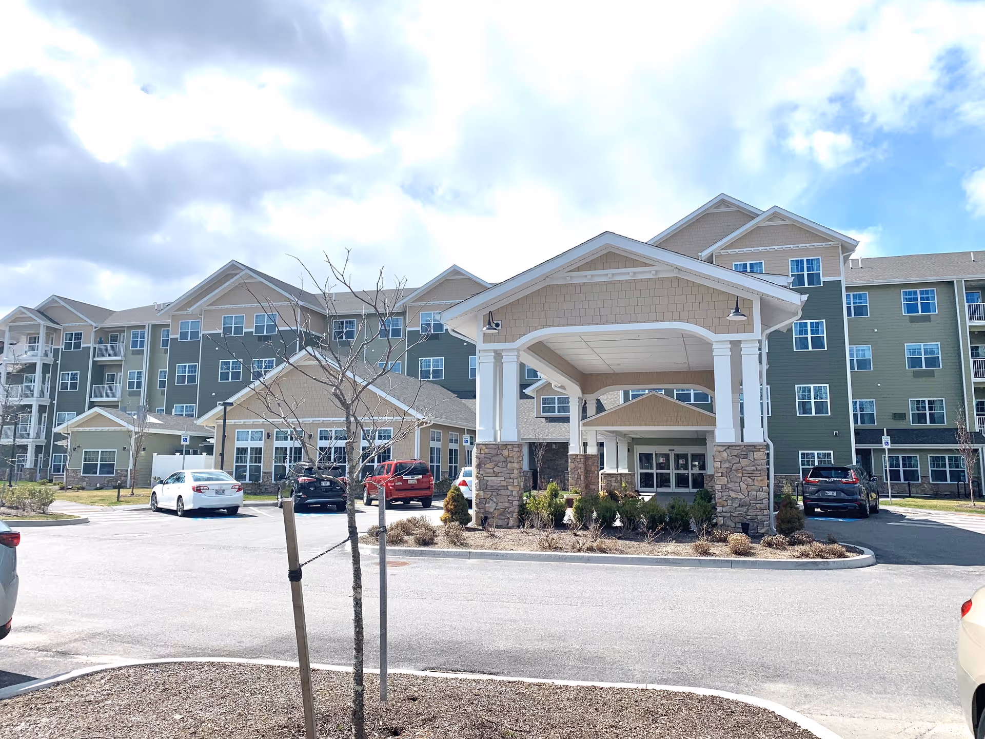 Front exterior view of Ashton Gardens Gracious Retirement Living facility showing a large multi-story building with green and beige siding, multiple windows, a covered entrance with stone pillars, several parked cars, and a partly cloudy sky.