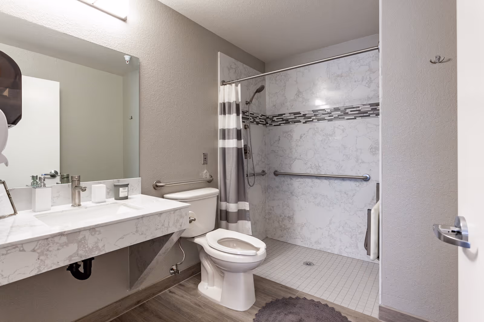 A modern bathroom with a white marble countertop sink, a toilet, and a walk-in shower with a gray and white striped shower curtain. The shower area has a tiled floor and walls with a decorative horizontal tile strip. There are grab bars installed near the toilet and inside the shower for accessibility. The floor is wood-style laminate, and a round gray bath mat is placed near the shower entrance.