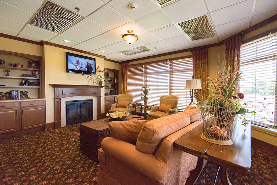 A cozy living room area with a large window covered by blinds and curtains, two armchairs, a sofa, a wooden coffee table, a fireplace with a mounted TV above it, built-in bookshelves, and decorative plants and lamps.