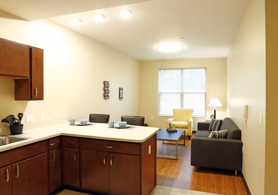 Interior view of a senior living apartment showing a kitchen area with wooden cabinets and a countertop set with dishes, leading into a living room with a gray sofa, yellow armchair, coffee table, floor lamp, and a window with blinds.