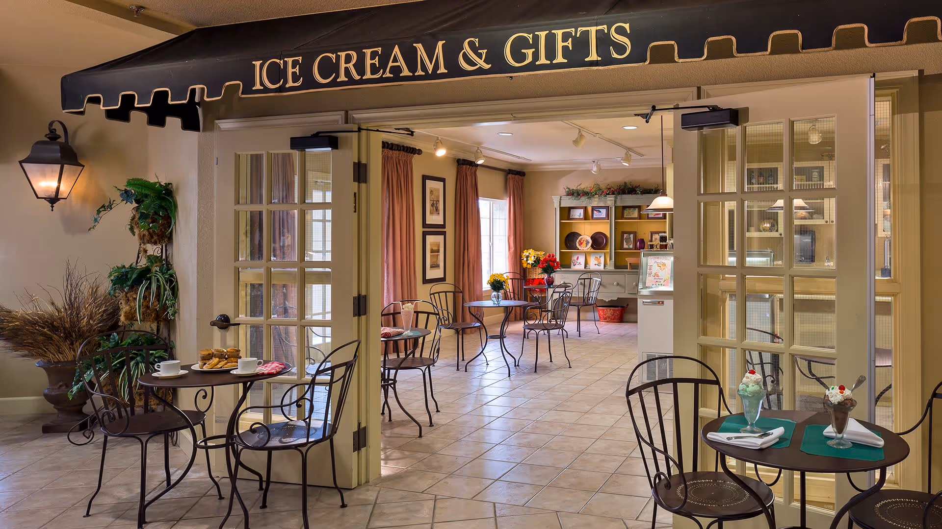 Interior view of a cozy ice cream and gifts shop area with small round tables and wrought iron chairs. The space has tiled floors, large windows with curtains, and decorative plants. Two tables near the entrance have ice cream sundaes and coffee cups placed on them. The shop sign above the entrance reads 'ICE CREAM & GIFTS'.