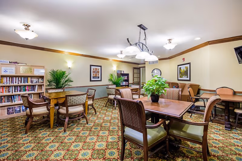 A well-lit common area with multiple wooden tables and cushioned chairs arranged for seating. The room features patterned carpet flooring, framed artwork on the walls, potted plants on tables and shelves, and a bookshelf filled with books. Ceiling lights and wall sconces provide illumination.