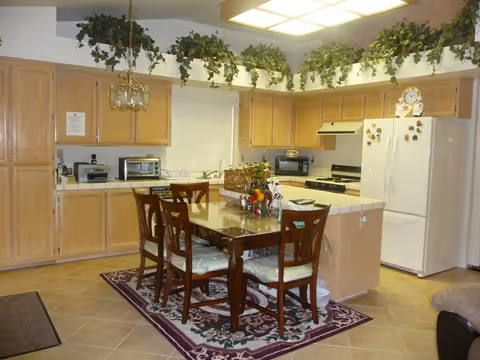 A kitchen with light wood cabinets, a white refrigerator, microwave ovens, and a stove. There is a dining table with six chairs on a patterned rug in the center. Green leafy plants are placed on top of the cabinets near the ceiling. The room is well-lit with ceiling lights.
