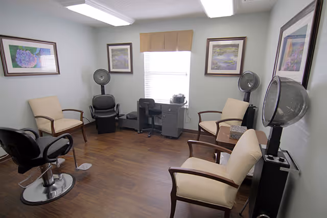 A small salon room with wooden flooring, featuring two hair drying chairs with hooded dryers, three beige armchairs, a black salon chair, a small desk with a chair, and framed artwork on the walls. A window with a beige valance allows natural light into the room.