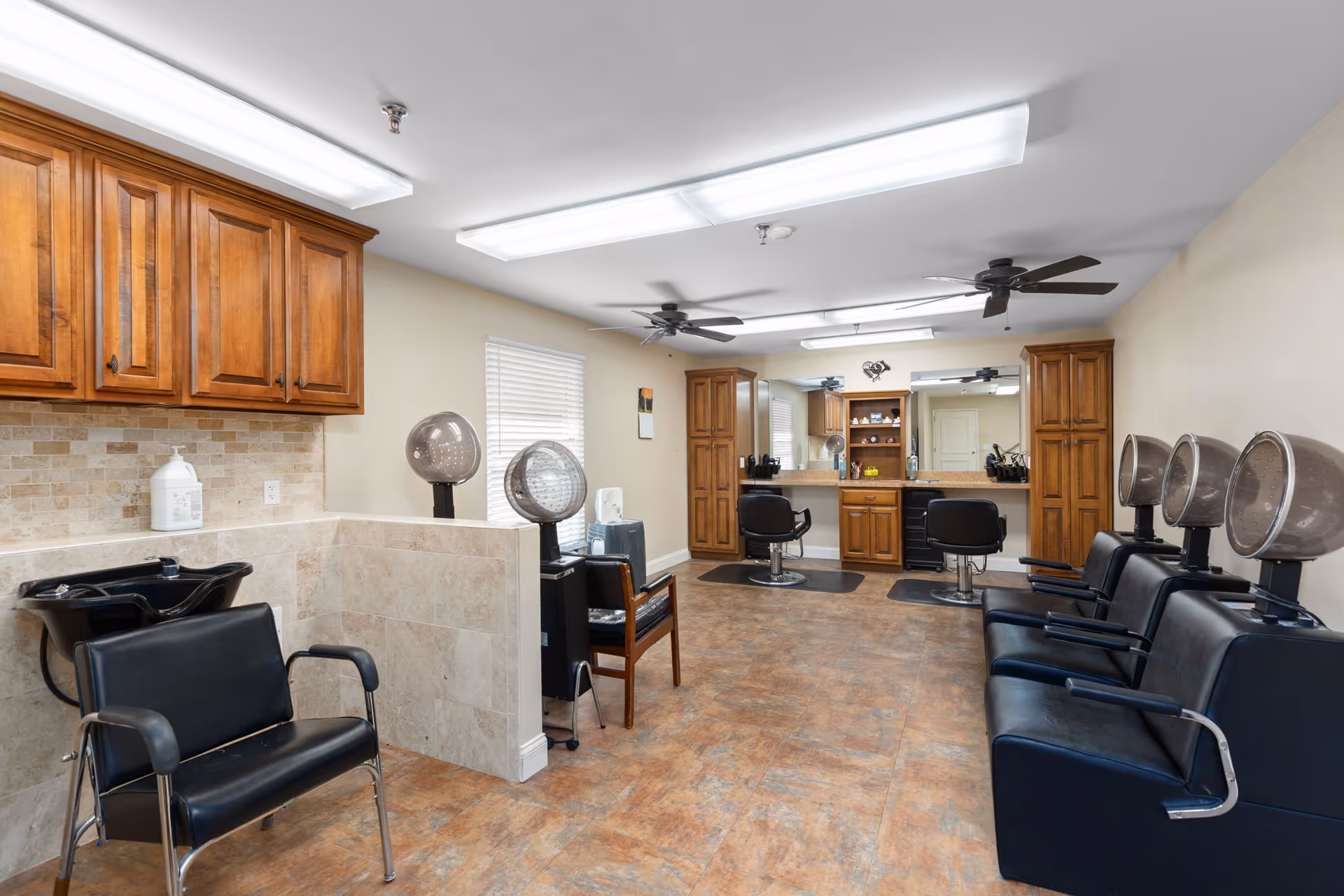 Interior view of a hair salon area in a senior living facility with multiple black salon chairs, hair dryers, wooden cabinets, and large mirrors on the wall. The room has beige walls, tiled flooring, and ceiling fans.