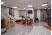 Interior view of a senior living facility common area featuring a small dining table with four chairs, a wall-mounted television above a fireplace, a daybed with yellow pillows, a staircase with wooden handrails, and a kitchenette with stainless steel appliances.