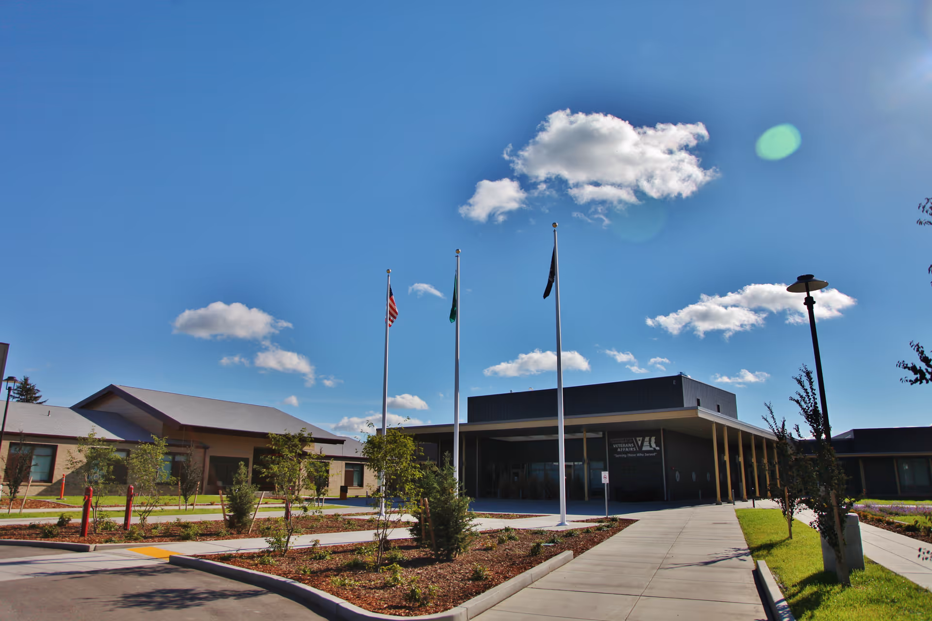 Exterior view of the Walla Walla Veterans Home building under a blue sky with scattered clouds. The building has a modern design with a covered entrance supported by columns. Three flagpoles with flags stand in front of the building, surrounded by landscaped greenery and a paved walkway.
