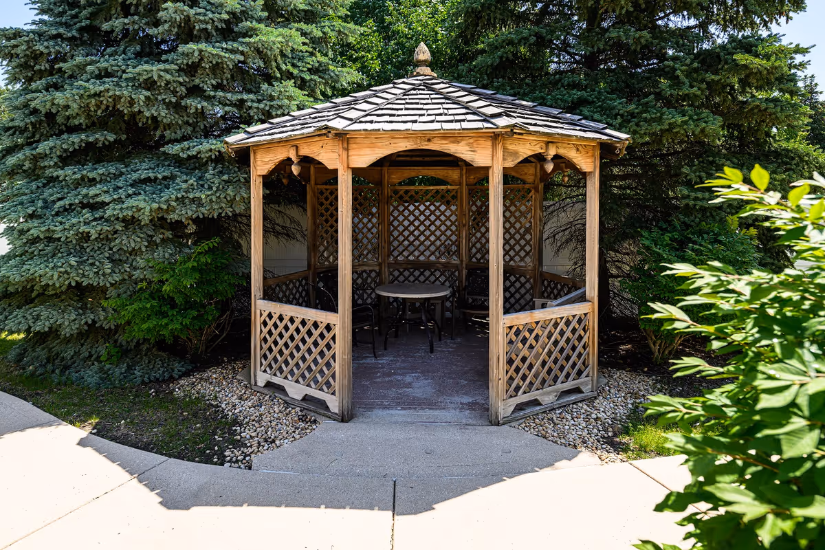 Wooden gazebo with a table and chairs nestled among evergreen trees in an outdoor garden.