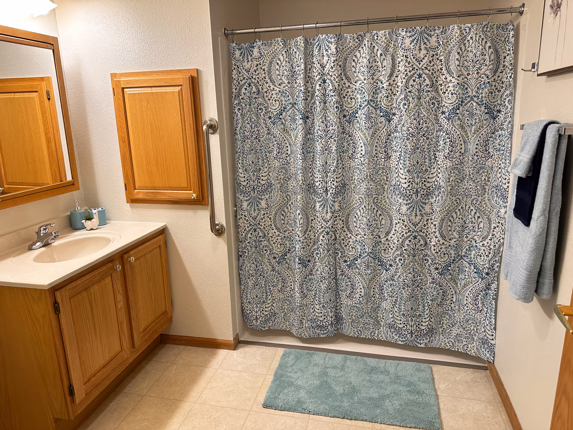 A bathroom with a wooden vanity cabinet and sink on the left, a wall-mounted wooden cabinet above it, a patterned shower curtain with blue and green designs, a silver grab bar next to the shower, a light blue bath mat on the tiled floor, and two towels hanging on a towel rack on the right wall.