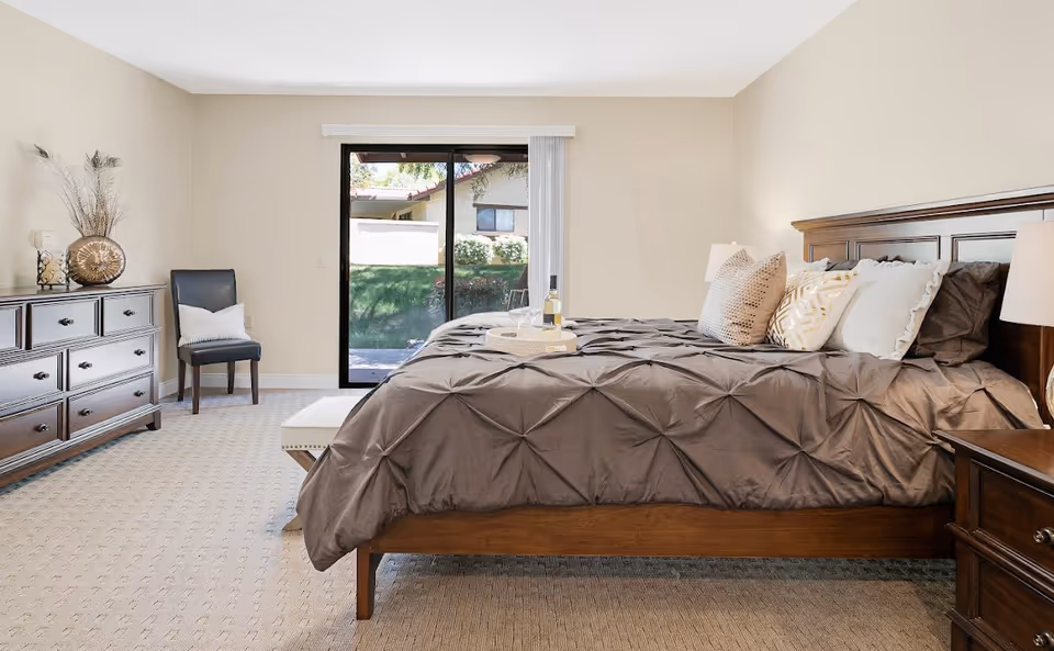 A spacious bedroom in HarborView Senior Assisted Living featuring a large wooden bed with gray bedding and multiple decorative pillows. There is a wooden dresser with decorative items on the left side, a dark chair with a white pillow next to it, and a sliding glass door leading to an outdoor area with greenery. The room has beige walls and carpeted flooring.