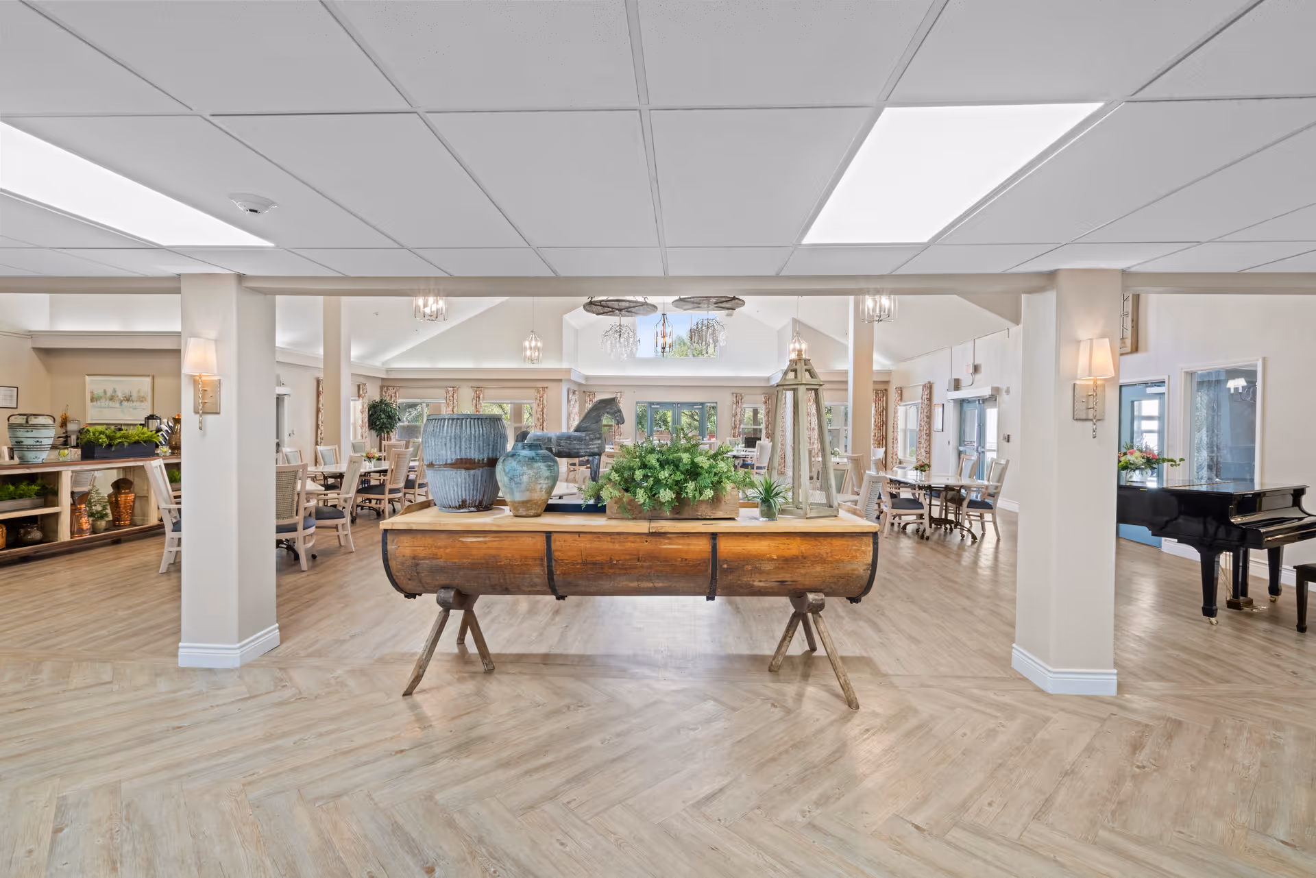 Spacious communal dining room with a central wooden display table holding plants and pottery, surrounded by dining tables and a piano.