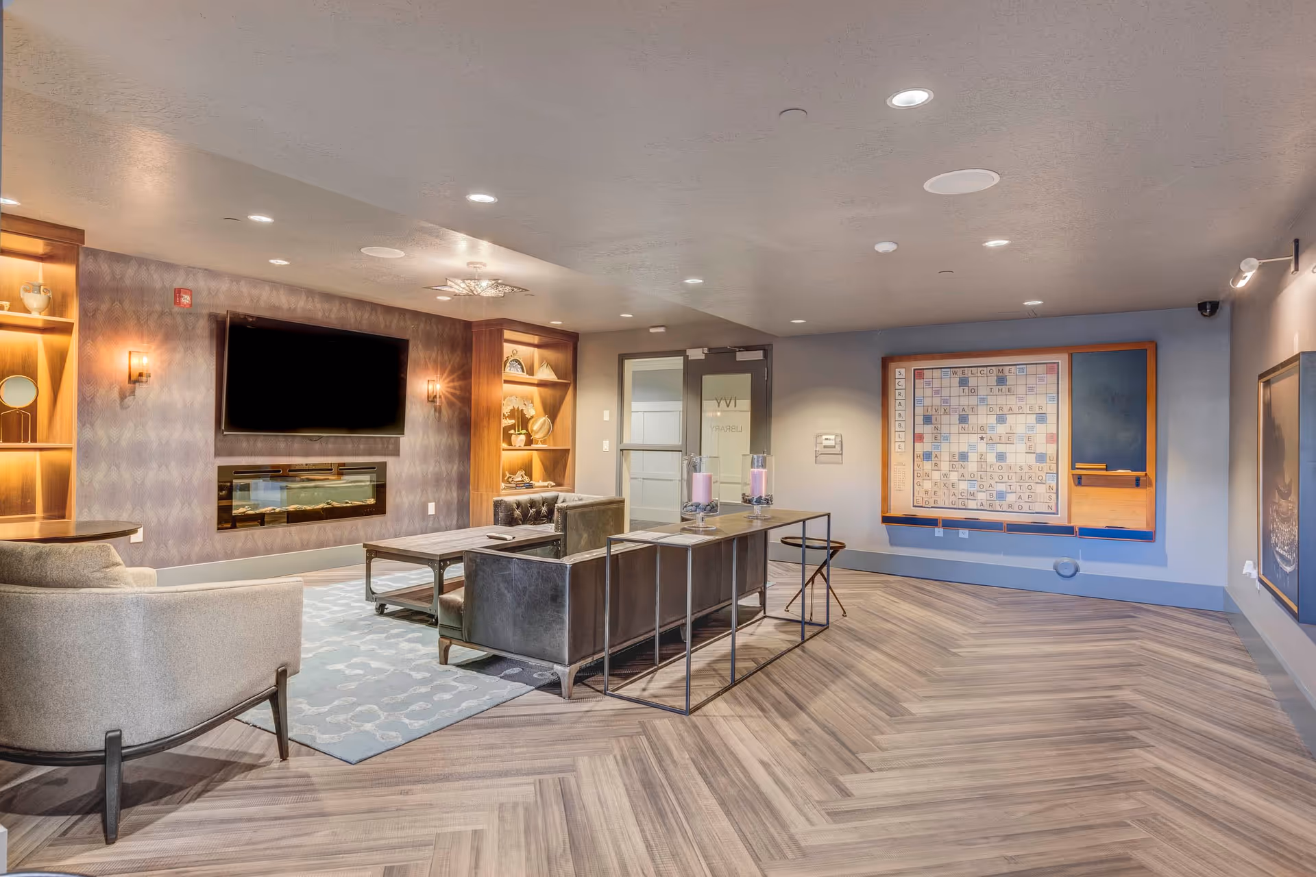A modern lounge area with a large flat-screen TV mounted above a sleek electric fireplace. The room features comfortable seating including a gray armchair and a leather sofa, a coffee table, and a console table with decorative candles. Built-in wooden shelves with decorative items flank the fireplace. On the far wall, there is a large Scrabble board with words welcoming visitors to The Ivy at Draper. The floor has a herringbone wood pattern, and the ceiling is fitted with recessed lighting.