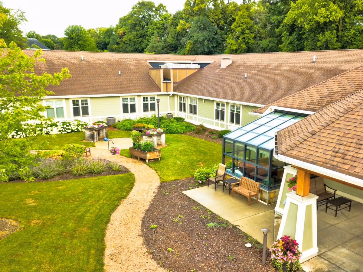 A landscaped outdoor courtyard area of a senior living facility with a winding gravel path, raised garden beds with flowers and plants, green grass, and a building with large windows and a glass sunroom. There are benches and chairs placed on a concrete patio area near the building, surrounded by trees and greenery.