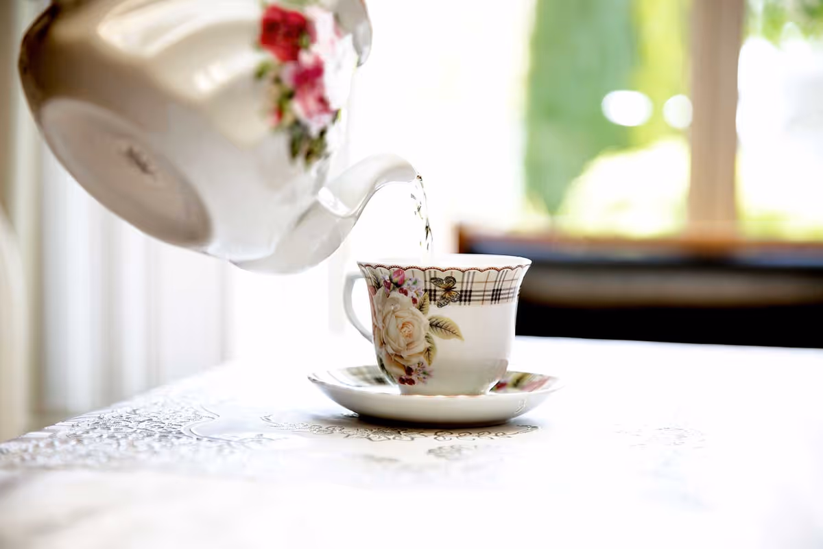 A close-up of a floral-patterned teapot pouring hot water into a matching teacup on a saucer, placed on a white lace tablecloth with a softly blurred background.