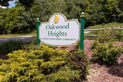 A green and white sign for Oakwood Heights, a Presbyterian Homes community, surrounded by bushes and trees with a road in the background.