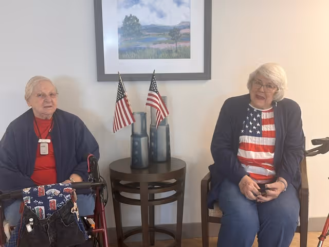 Two elderly women sitting indoors on chairs with a small round table between them holding two small American flags. A framed landscape painting hangs on the wall behind them. One woman is wearing a red shirt and dark jacket, the other is wearing a shirt with an American flag design and a dark jacket.