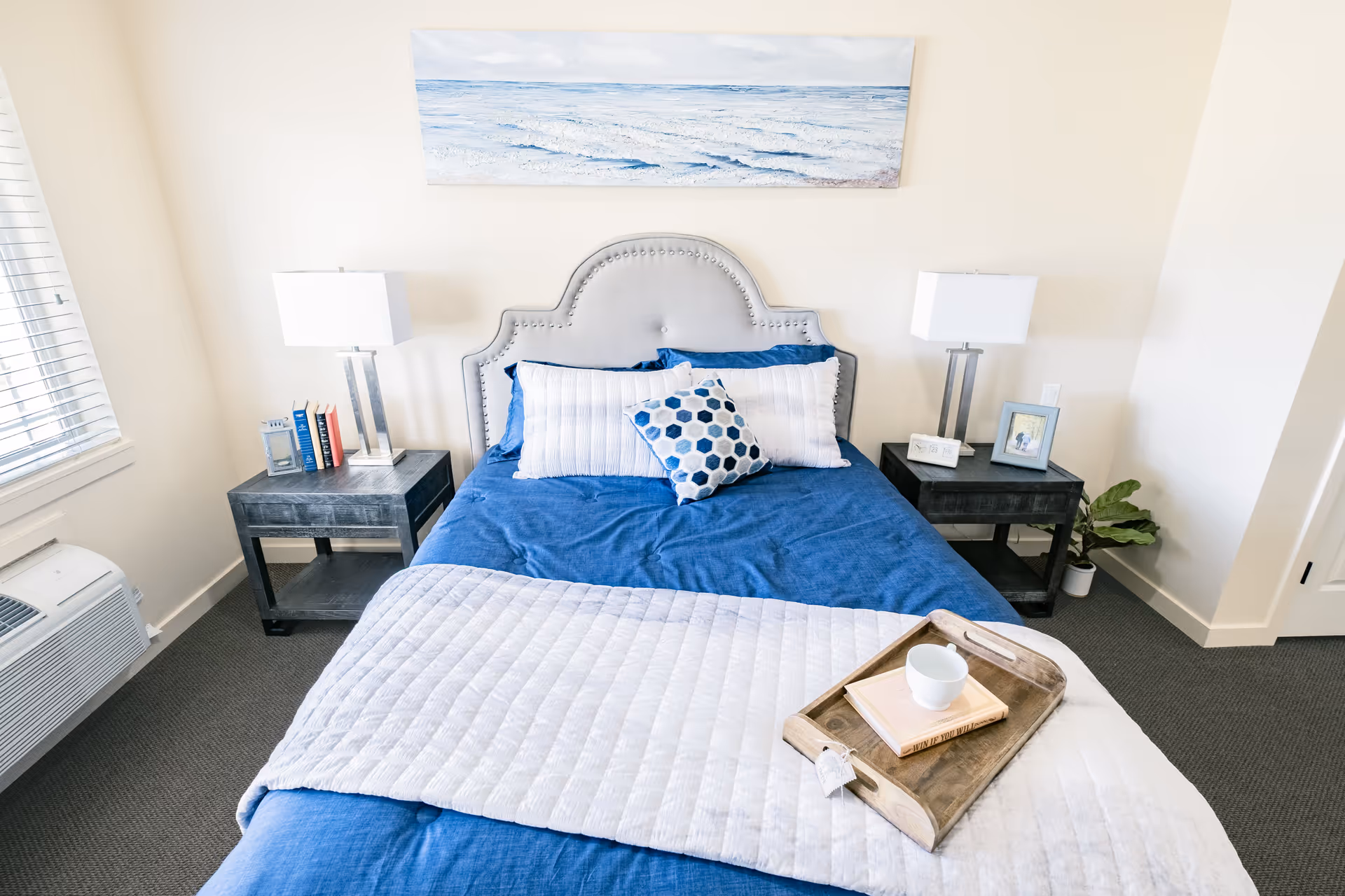 Bright bedroom featuring a bed with blue bedding, matching nightstands and lamps, wall art, and a tray with a cup on the bed.