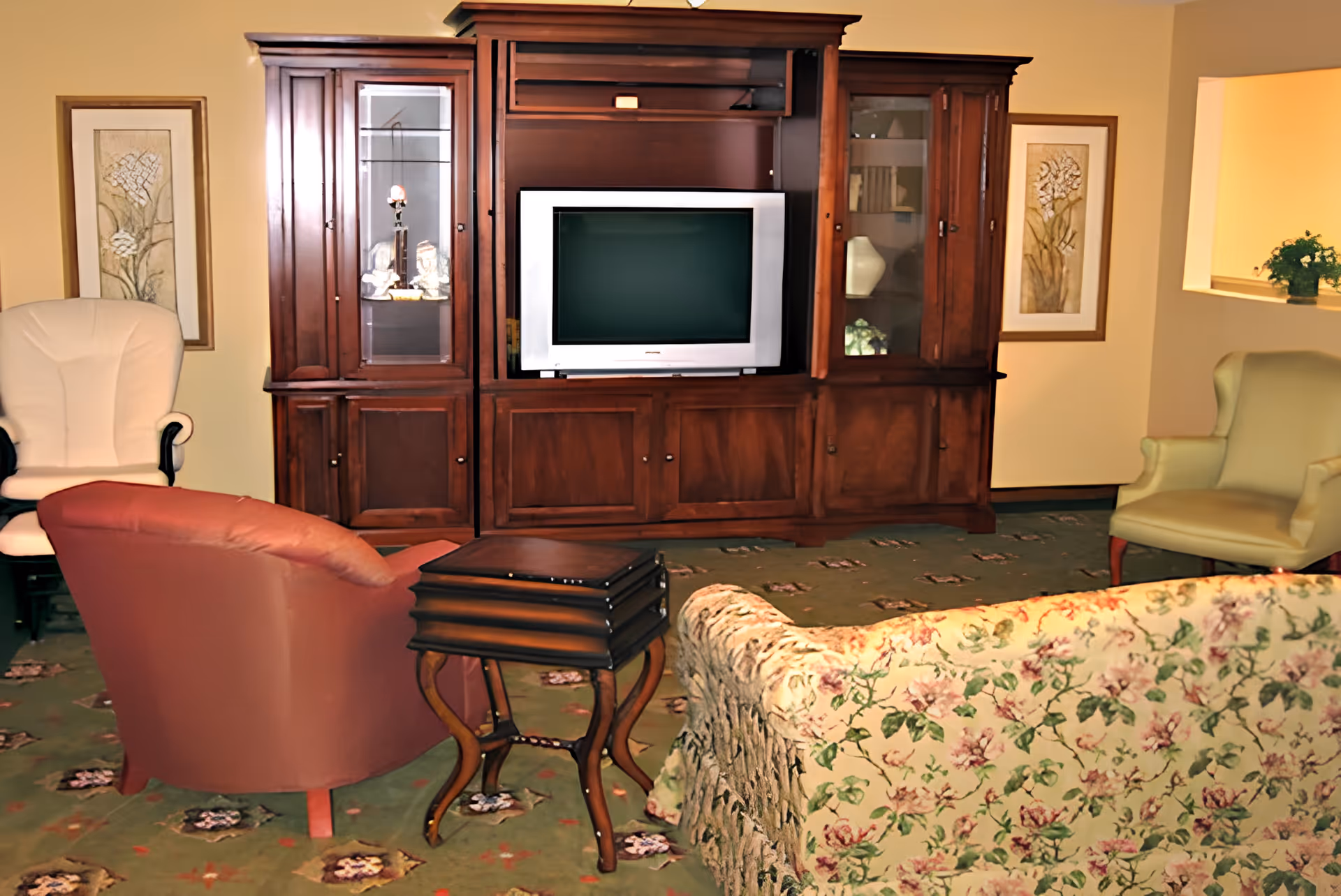 Living room with a large wooden entertainment center and television, several upholstered chairs, a floral sofa, and small nesting tables.