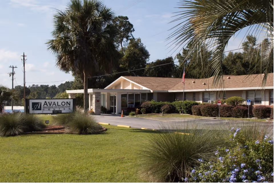 Exterior view of Avalon Healthcare and Rehabilitation Center building with a sign in front, surrounded by green grass, palm trees, and bushes under a clear sky.