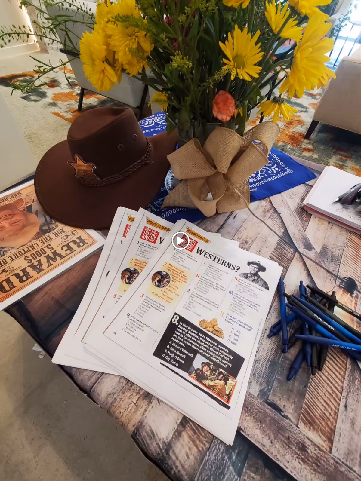 A table with a bouquet of yellow and orange flowers in a vase tied with a burlap bow, a brown cowboy hat with a sheriff star badge, several printed quiz sheets titled 'WESTERNS?', a stack of blue pens, and a newspaper with a 'REWARD' poster featuring a man's face.