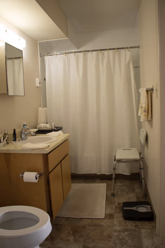 A bathroom with a white toilet, a wooden vanity with a sink and various toiletries, a mirror above the sink, a white shower curtain on a rod, a small white shower chair, a beige bath mat on the tiled floor, and a black bathroom scale near the chair.