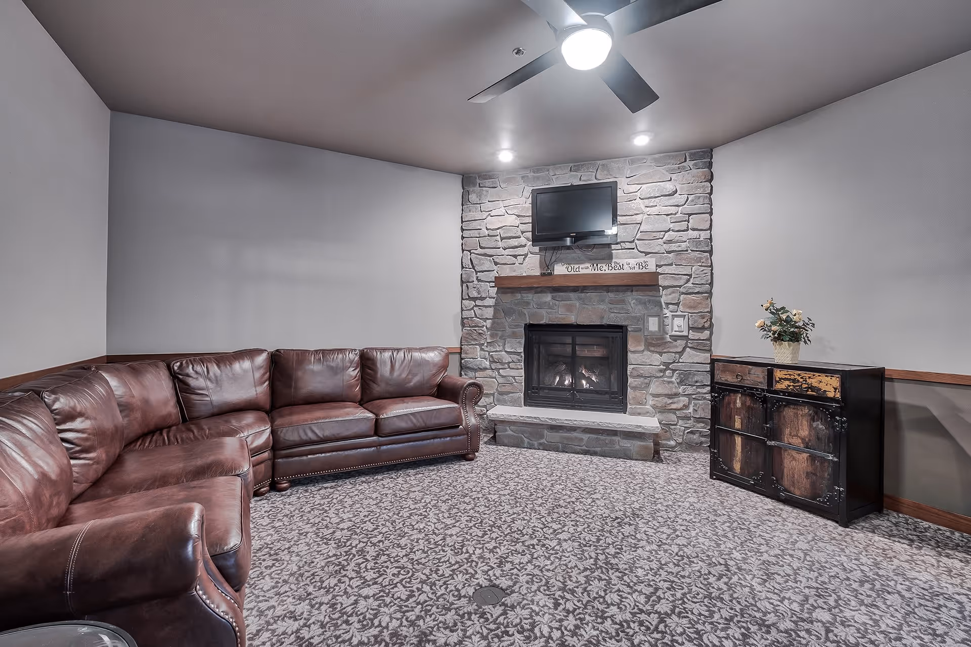 A cozy living room with a large brown leather sectional sofa, a stone fireplace with a wooden mantle, a flat-screen TV mounted above the fireplace, and a small cabinet with a flower arrangement on top. The room has patterned carpet flooring and neutral-colored walls.