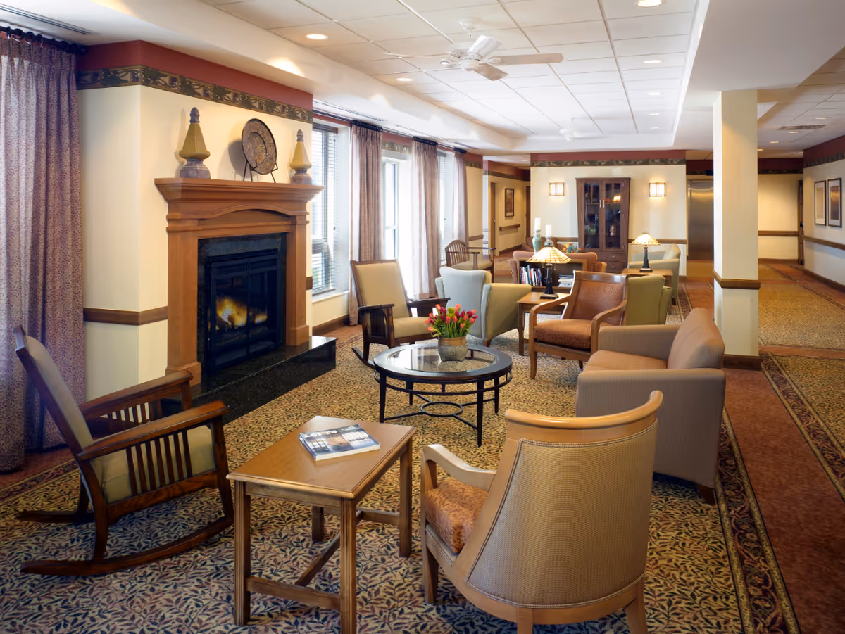 A cozy living room area in a senior living facility with multiple armchairs and a rocking chair arranged around a glass coffee table with a flower pot. There is a wooden fireplace with decorative items on the mantel, large windows with curtains, and warm lighting from table lamps and wall sconces. The room has patterned carpet and a ceiling fan.