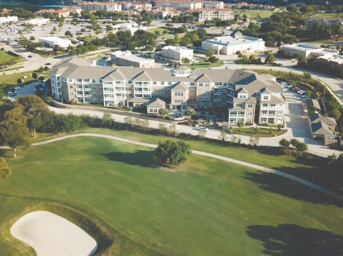 Aerial view of a large multi-story residential building surrounded by greenery, with a golf course and sand trap in the foreground. The building has multiple balconies and is situated in a suburban area with other buildings and roads visible in the background.