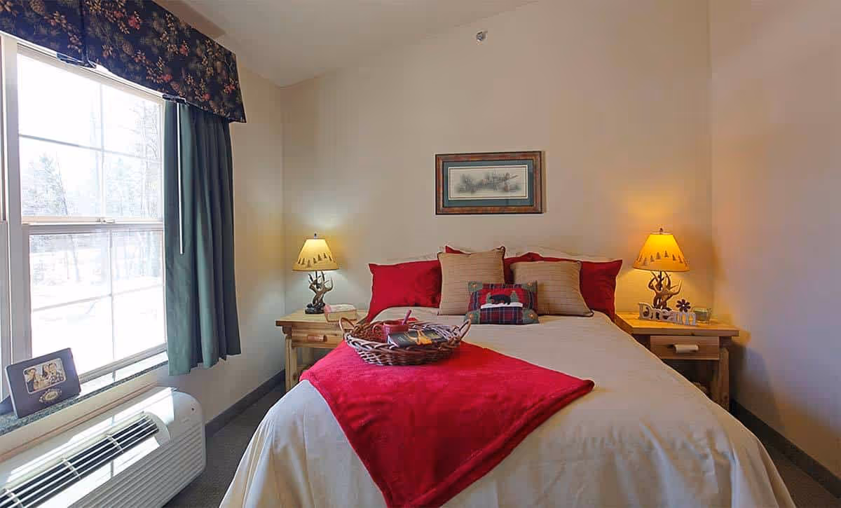 A cozy bedroom with a made bed featuring red accents, bedside tables with lamps, and a window with curtains.