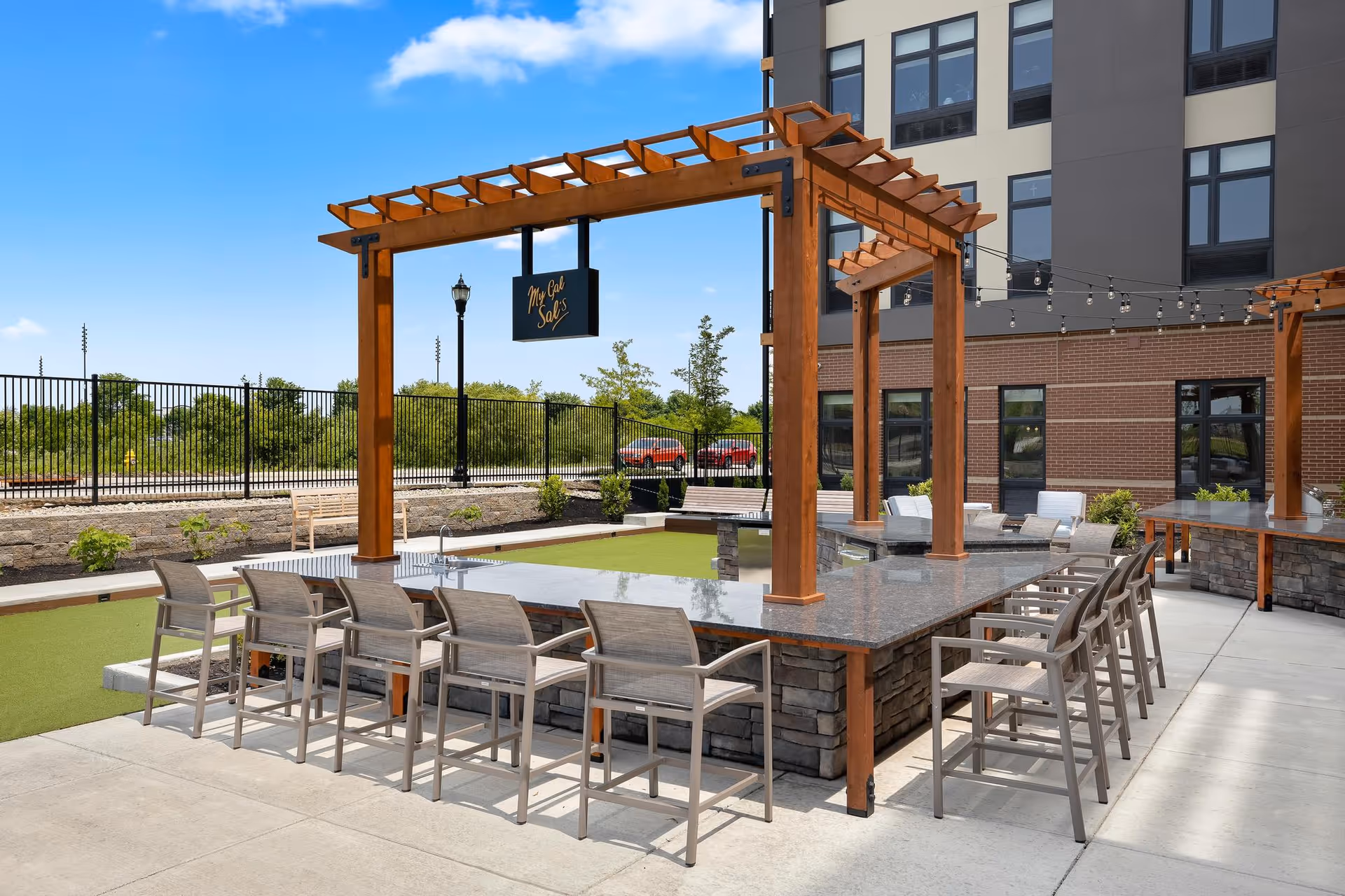 Outdoor seating area with a large rectangular stone and granite countertop surrounded by multiple chairs under a wooden pergola structure. The area is part of a senior living facility with a building in the background, string lights hanging, and a sign that reads 'Mr. Cal Sal's'. There is a fenced green space and clear blue sky.